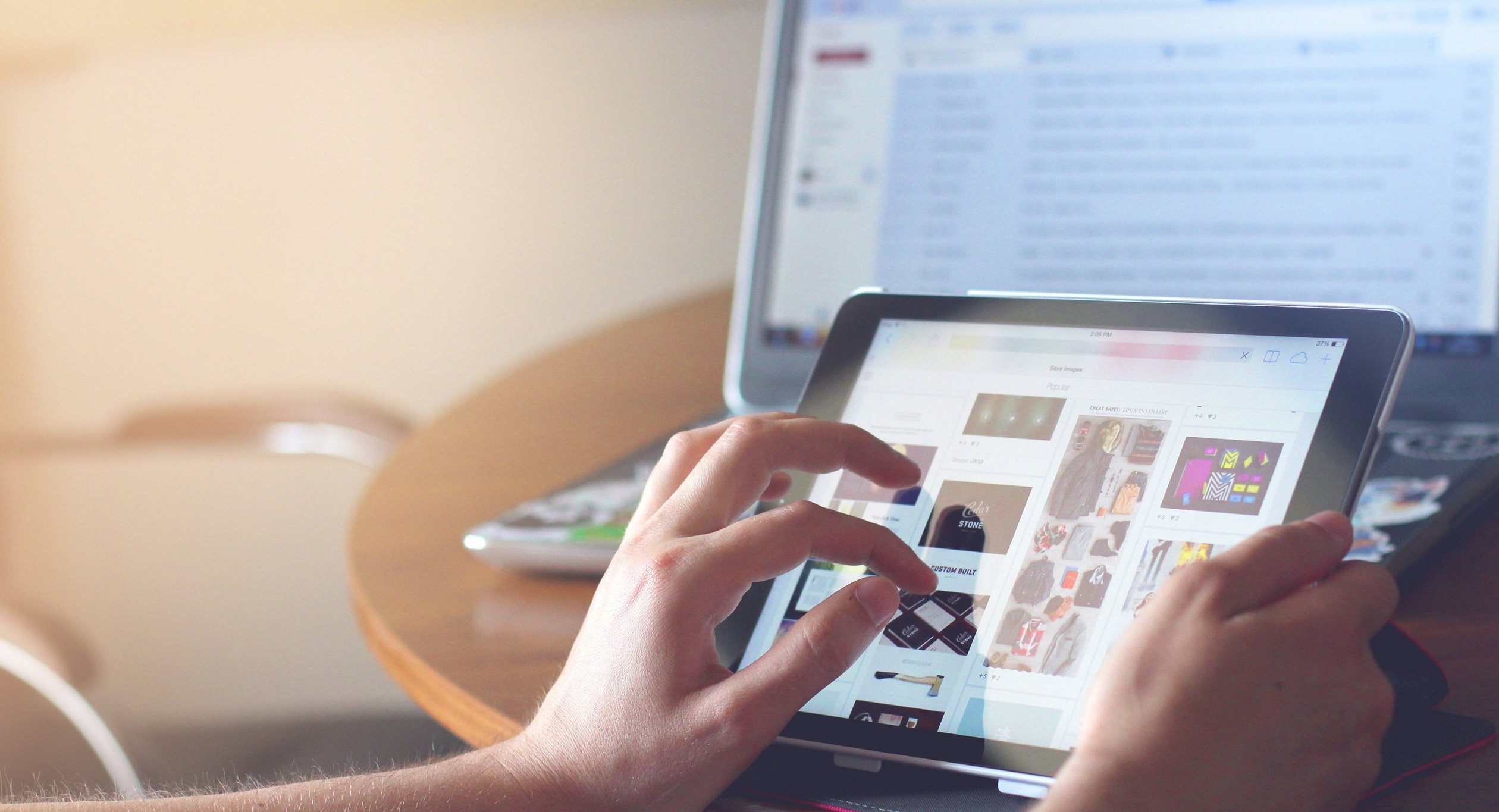Hands hold an iPad while a laptop screen behind it shows email open.