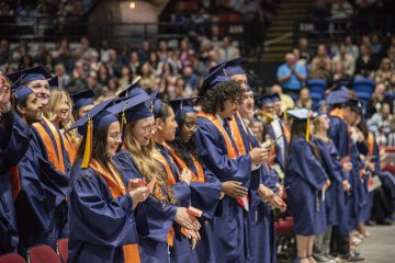 A row of graduates in blue commencement gowns and caps in an auditorium.