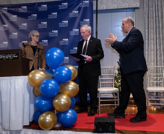 Dr. Thomas Rossi stands center on a platform stage, holding a plaque, as Provost Stephanie Nesbitt stands left and Genesis Group head Ray Durso to the right, both clapping.