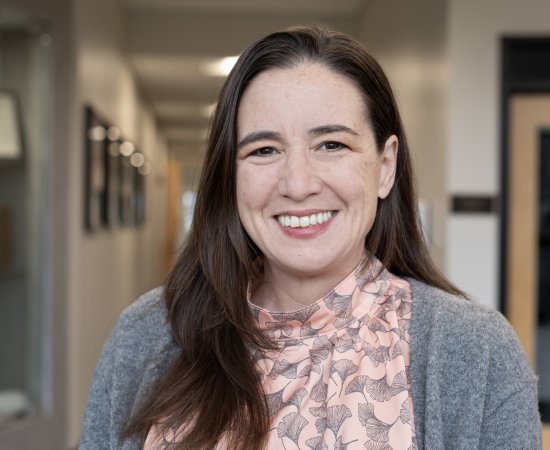 Deborah Pollack, with long brown hair, pink top and gray sweater, smiles at camera.