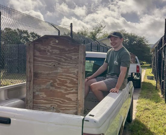 Marshall Hildreth in shorts, green t-shirt, and hat, sits in the back of a pick-up truck.