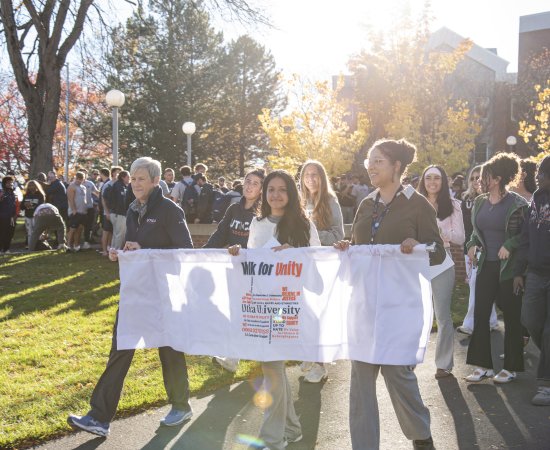President Nesbitt, student Madison Jordan, and Director of Community, Belonging and Engagement Kira Maddox carry a banner as a crowd of students, faculty, and staff follow behind them.