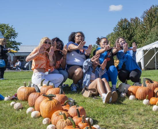 Students sit at the top of a big U made out of pumpkins on the grass of campus, making a U with their hands.