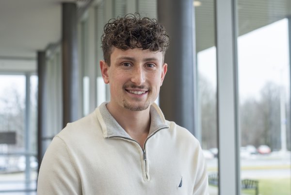 Conner Crossley, in white zip-up pullover, stands by a window in Bull Hall and smiles.