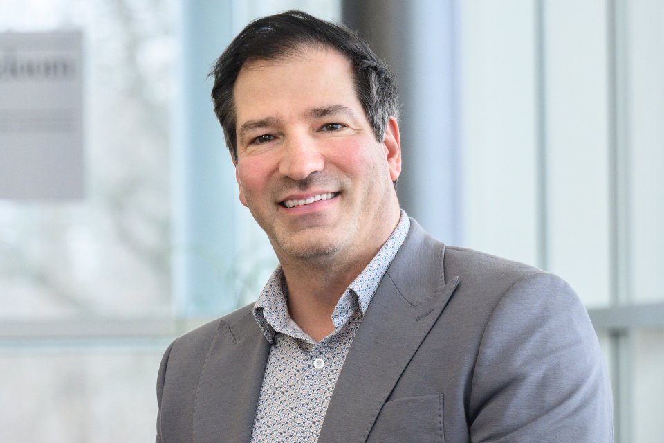 Dr. Christopher Riddle in gray suit jacket sits in front of a wall of windows and smiles.